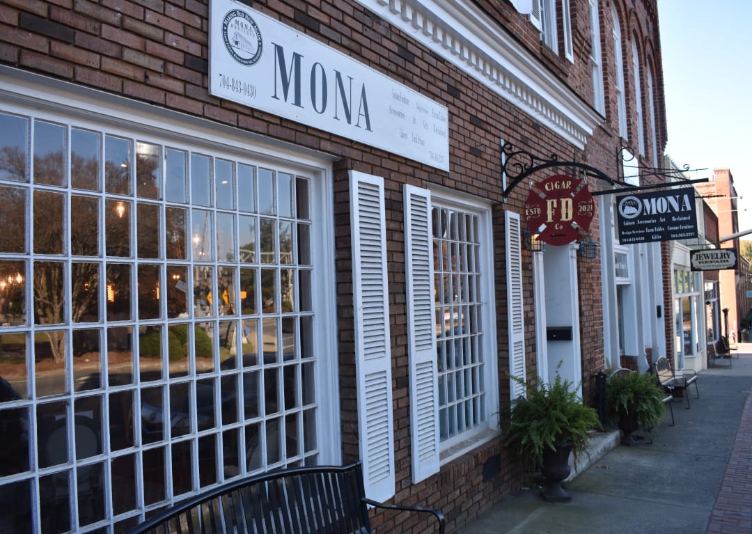 The MONA Boutique storefront on East South Main Street in Waxhaw, NC, with white-trimmed windows and hanging signs on a brick building